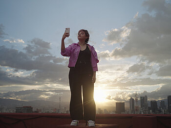 Eine junge Frau steht mittig im Bild und macht ein Selfie vor der Skyline von Mexiko City im Bildhintergrund. Der Himmel ist von Wolken zerklüftet, die Sonne geht gerade auf.