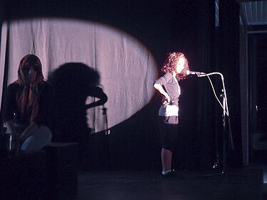 Bühnenszene mit zwei Jugendlichen: eine im Scheinwerferlicht, die andere im Schatten.