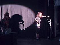 Bühnenszene mit zwei Jugendlichen: eine im Scheinwerferlicht, die andere im Schatten.