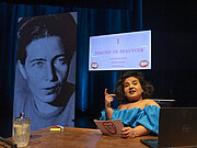 Eine Frau in blauem Kleid sitzt vor einem Laptop. Sie hat eine Moderationskarte in der Hand und hinter ihr auf einem Bildschrim steht der Name "Simone de Beauvoir".