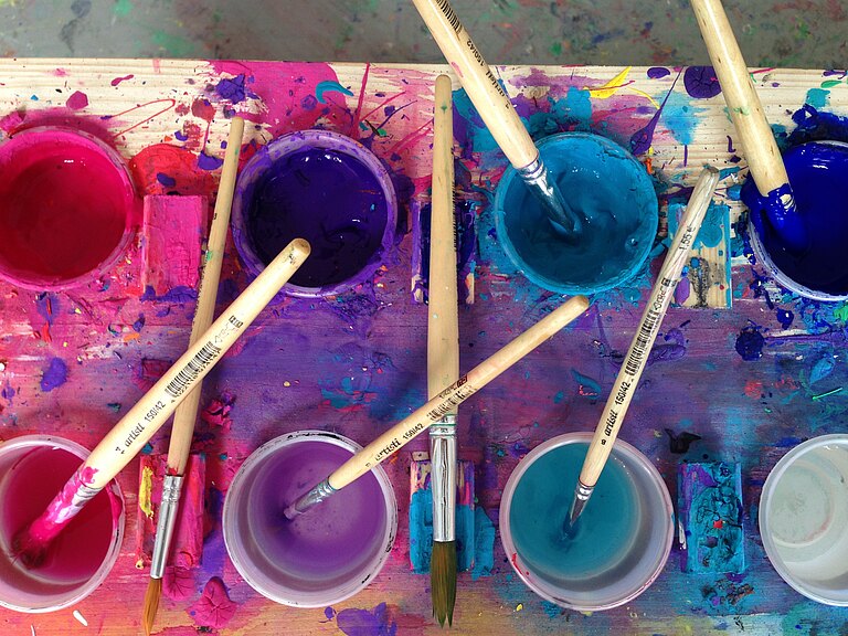 Das Atelier Artig benutzt besondere Farben für sein kunsttherapeutisches Arbeiten. Hier ist ein großer Farbkasten zu sehen mit Töpfen voll mit Nachtblau, Mittelblau, Türkis, Pink, Violett und Dunkellila.