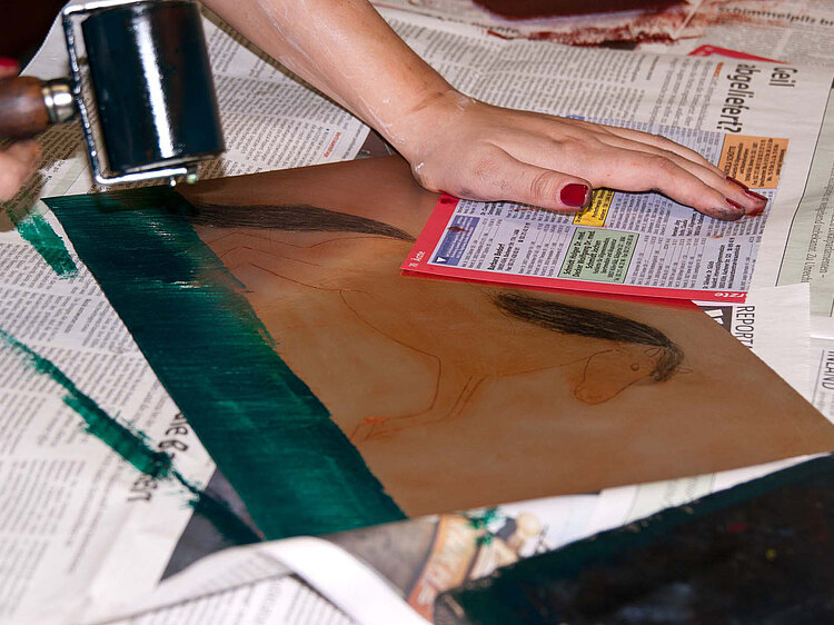 Auf dem Tisch der Druckwerkstatt walzt eine Frauenhand eine Platte mit Pferdegravur grün ein.