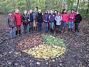 Landschaftskunst: Ein Kreis aus verschiedenfarbigen Blättern liegt auf dem Boden vor einer Gruppe Jugendlicher im Wald.