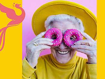 Bild für unseren Theater Spielclub für Menschen ab 60 Jahren. Eine fröhlich lächelnde ältere Dame hält sich zwei pinke Donuts vor die Augen. Sie trägt einen gelben Hut und einen gelben Pullover. 