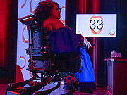 Eine Frau sitzt im Rollstuh und trägt ein blaues Kleid. Der Hintergrund ist rot-belichtet und man sieht einen Bildschirm mit einem Mund und einer "33"