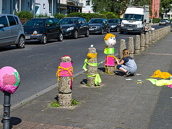 Straßenansicht, durch die diagonal eine Reihe Begrenzungspöller führt. Drei davon sind mit bunter Wolle gestaltet, an einem weiteren bastelt gerade ein Mädchen.
