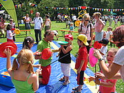 Große Veranstaltung im Park: Im Vordergrund leitet ein Teller-Jongleur ein kleines Mädchen an.