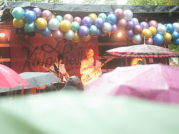 Blick auf die Bühne, geschmückt mit Luftballons, wir sehen Menschen die Schirme halten im Zuschauer:innen-Bereich
