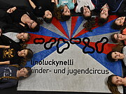 12 Kinder liegen strahlenförmig auf einem Teppich mit dem Aufdruck "linoluckynelli" Kinder- und Jugendzirkus.