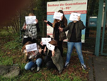 Eine Gruppe Jugendlicher hält sich Gesichterzeichnungen vor die Köpfe. Im Hintergrund das Eingangsschild des "Bauhaus der Generationen"