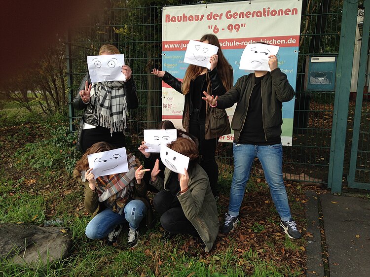 Eine Gruppe Jugendlicher hält sich Gesichterzeichnungen vor die Köpfe. Im Hintergrund das Eingangsschild des "Bauhaus der Generationen"