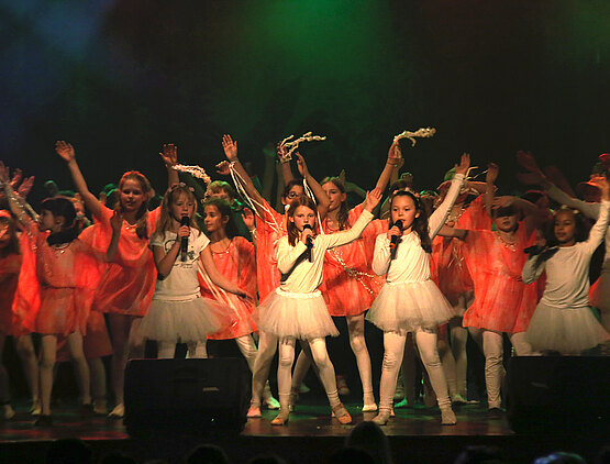 Eine grün beleuchtete Bühne voller winkender Kinder in weißen und flamingofarbenen Tanzkostümen.