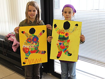 Zwei Mädchen halten große gelbe Plakate mit bunten "Nana"-Figuren in die Kamera.