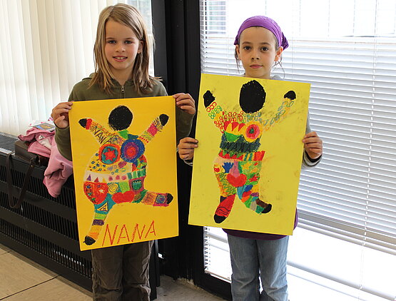 Zwei Mädchen halten große gelbe Plakate mit bunten "Nana"-Figuren in die Kamera.