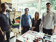 Vier Erwachsene stehen zwischen einer Staffelei und den Kreativmaterialien in einem hellen Raum
