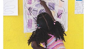 Ein Mädchen in Rückenansicht steht vor einer gelben Wand und zeigt auf ein Bild.