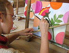 Sehr konzentriert benutzt ein Mädchen einen Malstock zum konturieren eines floralen Motivs auf einer bunten Leinwand.