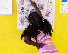 Ein Mädchen deutet ausdrucksvoll auf Elemente einer großformatigen Zeichnung in Lila-Tönen, die an einer gelben Wand hängt.