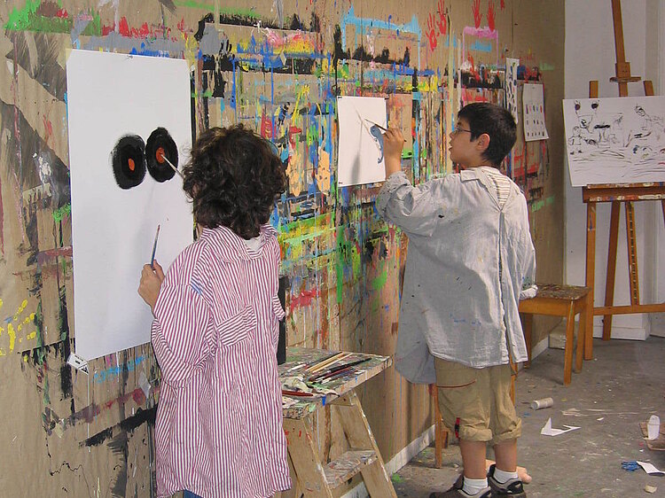 zwei Kinder arbeiten an einer großen Malwand voller Farbreste an ihren weißen Bögen