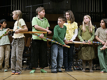 Sieben Kinder haben sich vor einer Orgel aufgestellt und halten stabartige, selbst gebastelte Instrumente.