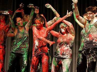 Eine Gruppe Jugendlicher tanzt im Stehen, über und über mit roter, grüner und weißer Farbe bemalt.