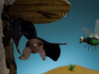 Filmstill aus Kopfüber: Eine Puppentrick-Fledermaus hägt kopfüber an einem Baumstamm; von rechts kommt eine Libelle ins Bild geflogen.