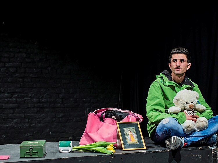 Bühnenszene aus Planet Heimat: Ein junger Mann mit Teddybär und kleinem Gepäck auf dem Bühnenrand sitzend.