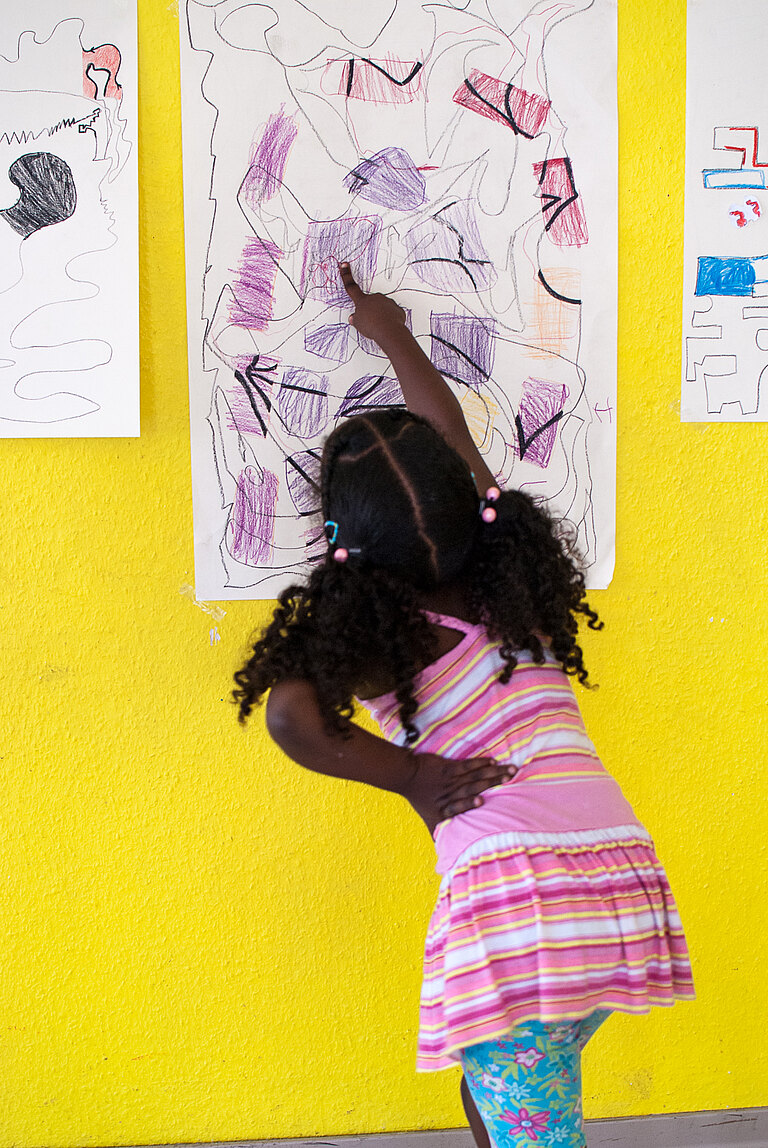 Ein Mädchen deutet ausdrucksvoll auf Elemente einer großformatigen Zeichnung in Lila-Tönen, die an einer gelben Wand hängt.  
