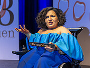 Eine Frau sitzt im Rollstuhl. Sie trägt ein blaues Kleid und hält einen Zollstock mit der Aufschrift "Sie kann's besser".