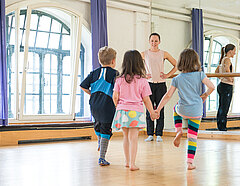 Kindertanz vor verspiegelter Wand: Eine Tanzlehrerin lächelt drei Kinder an, die auf sie zuspringen.